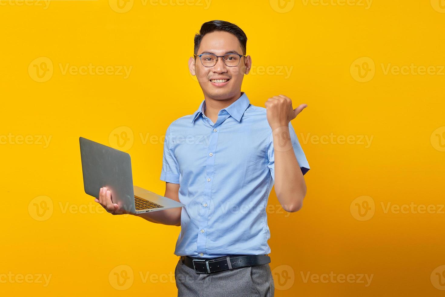 porträt eines gutaussehenden lächelnden asiatischen mannes mit brille, der einen laptop verwendet und mit dem finger auf den kopierraum zeigt, der auf gelbem hintergrund isoliert ist. geschäftsmann- und unternehmerkonzept foto