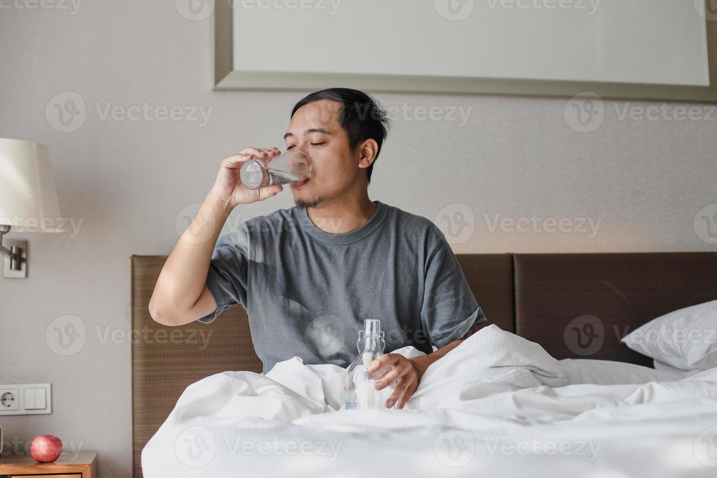 Mann sitzt auf dem Bett und trinkt ein Glas Mineralwasser nach dem Aufwachen aus dem Schlaf foto