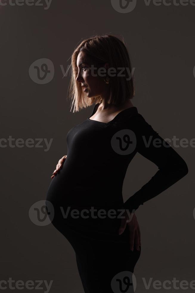stilvolle schöne schwangere frau in einem schwarzen kleid hält ihre hände auf ihrem bauch. foto