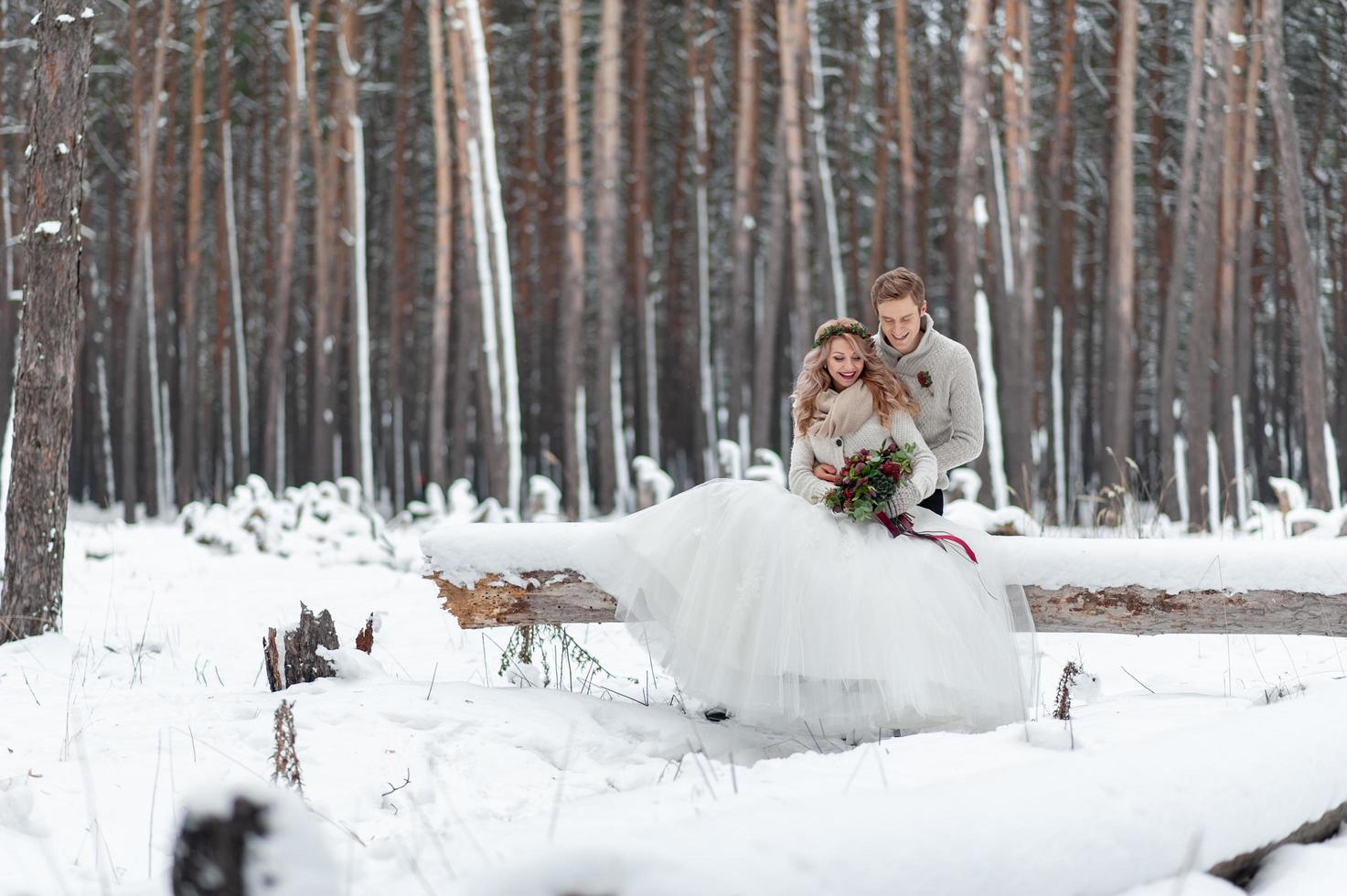 Süßes Paar, das sich in einen Blumenstrauß verliebt, sitzt auf dem Baumstamm im Hintergrund des Winterwaldes. Kunstwerk. Winterhochzeit. Platz kopieren foto