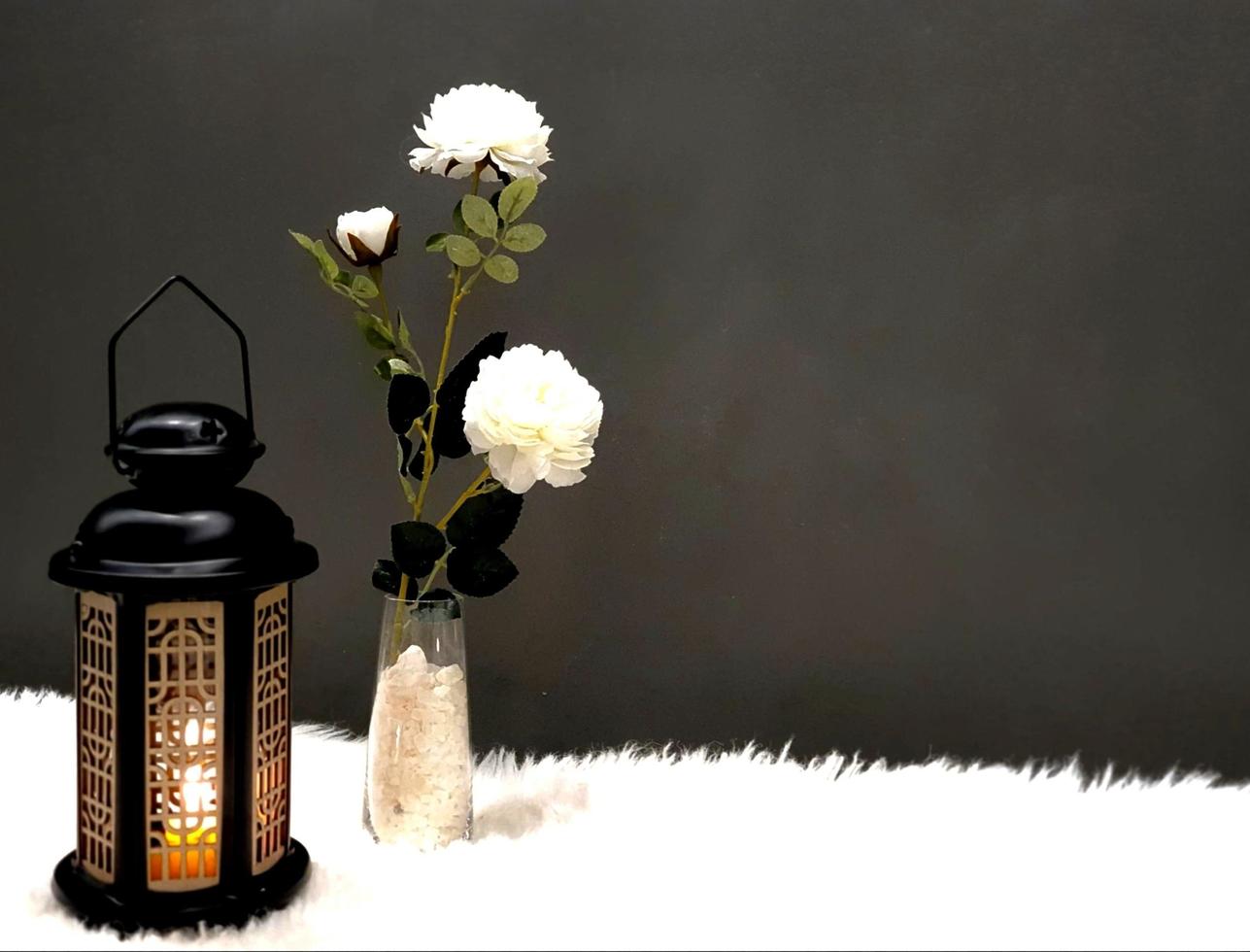 Die Ramadan-Laterne ist schwarz, leuchtend und ihre Seiten sind mit Holzdekorationen verziert, die neben einer kleinen Vase stehen, die weiße Kristalle enthält und auch einige weiße Rosen trägt, beides foto