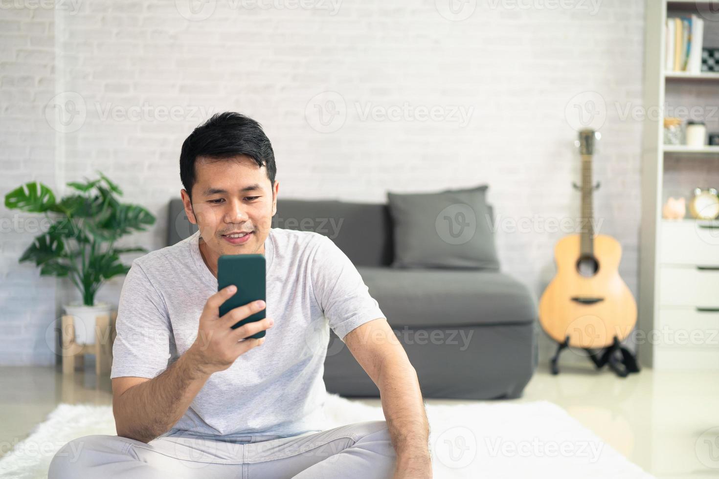 lächelnder asiatischer mann, der lässige kleidung trägt, die auf einer couch im wohnzimmer sitzt. asiatischer mann, der im wohnzimmer mit dem handy chattet. foto