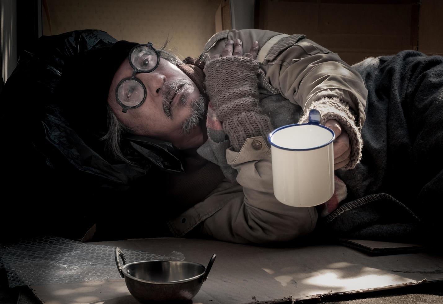 Alter Obdachloser mit grauem Bart liegt und hält Becher in der Hand und bettelt um Essen und Geld. foto