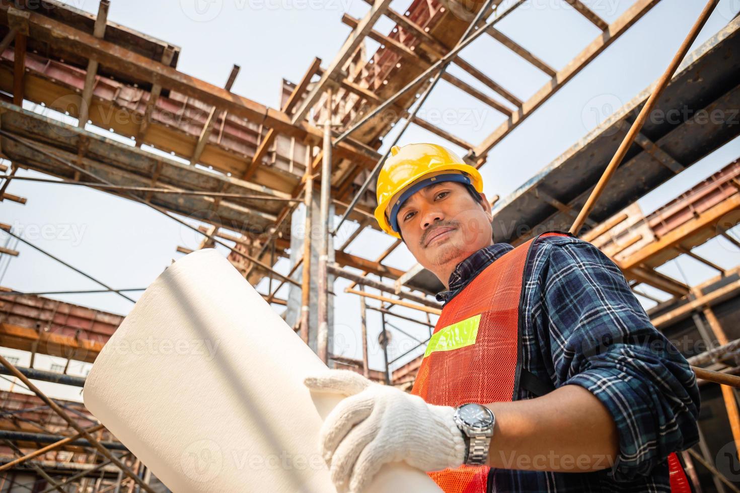 Arbeiter mit Schutzhelm, der eine Blaupause hält und auf die Kamera blickt, die das Projekt auf der Baustelle überprüft und plant foto