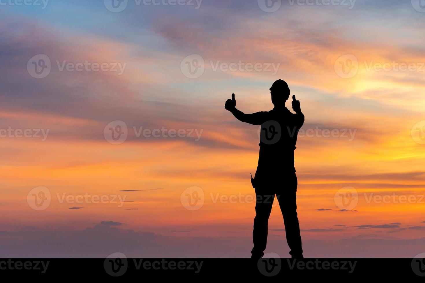 Silhouette des Fabrikarbeiters mit Beschneidungspfad im Schutzhelm mit Daumen nach oben, junger Mann feiert mit der Hand im Luftsonnenunterganghintergrund foto