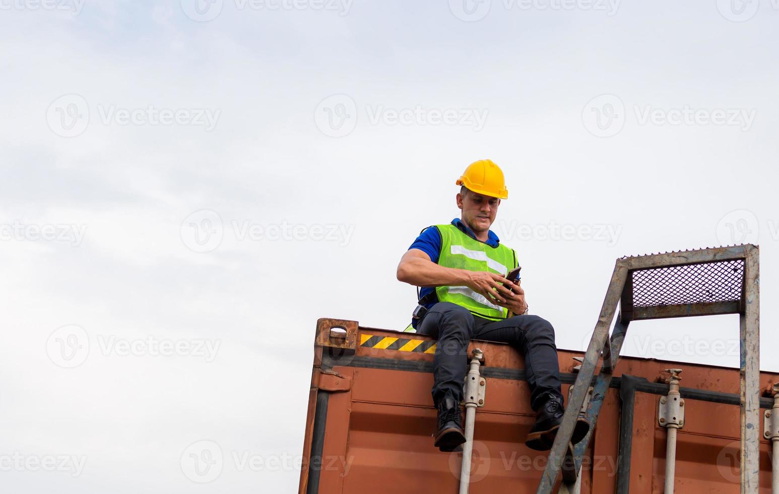 Vorarbeiter mit Schutzhelm und Sicherheitsweste, der auf einer Containerbox sitzt und auf das mobile Smartphone auf die Fracht blickt foto