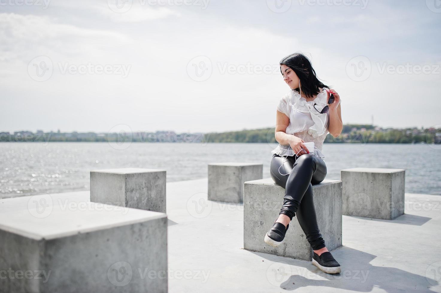 porträt eines brünetten mädchens, das musik über kopfhörer vom handy hört, lederhosen und weiße bluse für damen trägt, sonnenbrille, gegen steinwürfel am strand des sees. foto