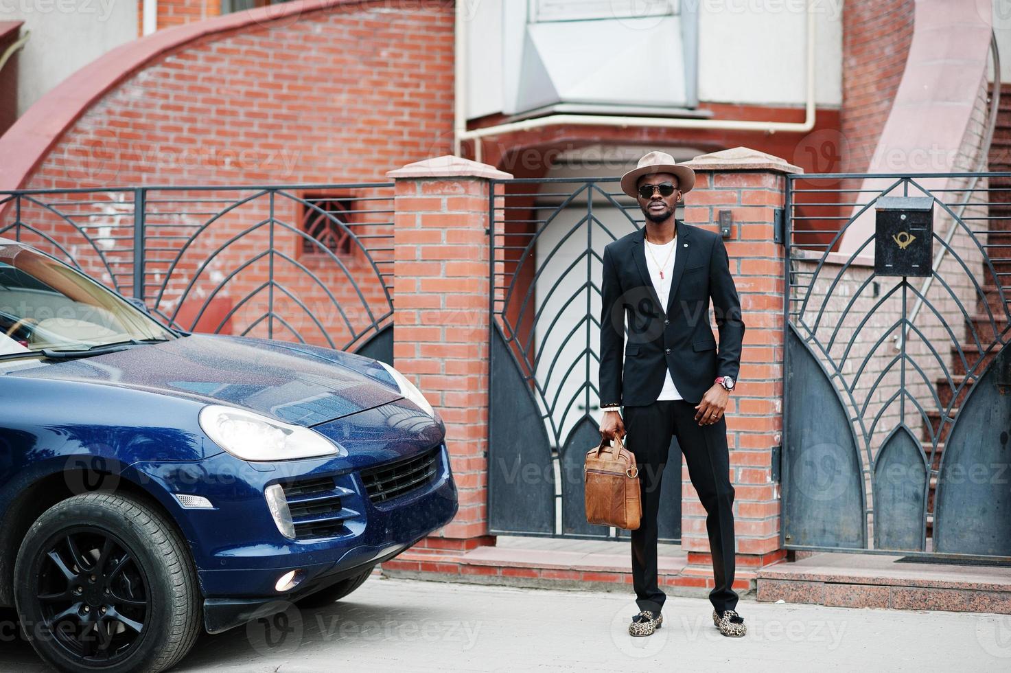 stilvoller schwarzer mann mit brille mit hut, anzug mit handtasche gegen luxusauto tragen. reicher afroamerikanischer geschäftsmann. foto