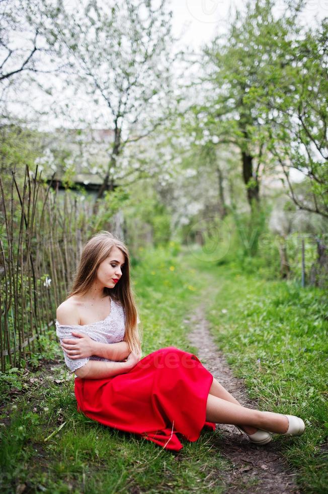 Porträt eines sitzenden schönen Mädchens mit roten Lippen im Frühlingsblütengarten auf grünem Gras, tragen rotes Kleid und weiße Bluse. foto