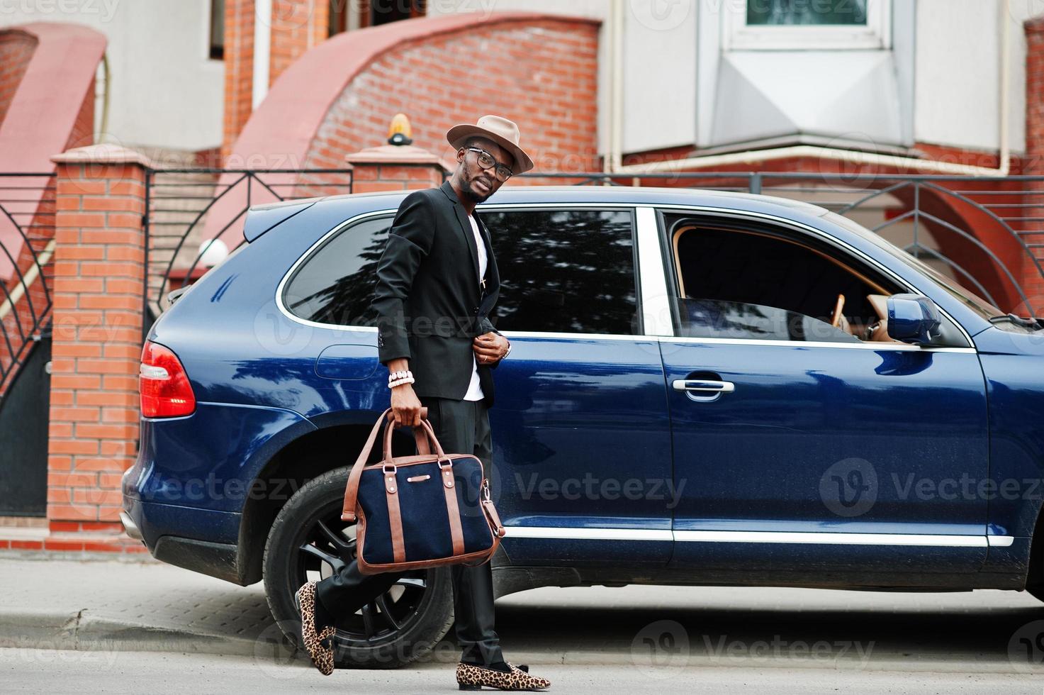 stilvoller schwarzer mann mit brille mit hut, anzug mit handtasche gegen luxusauto tragen. reicher afroamerikanischer geschäftsmann. foto