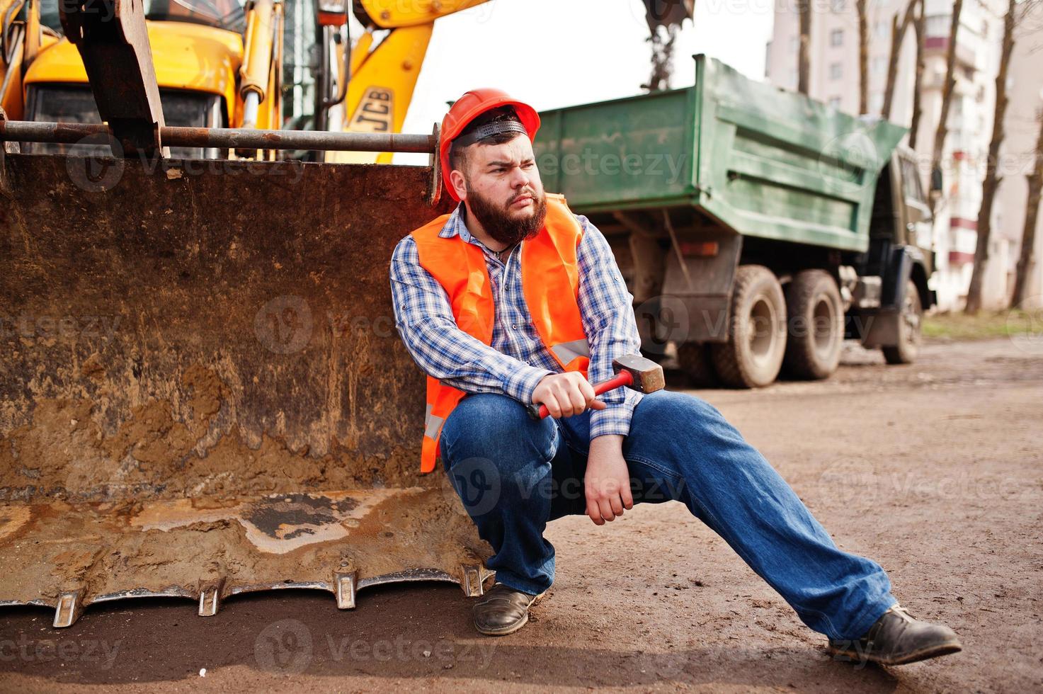 bart arbeiter mann anzug bauarbeiter in orangefarbenem sicherheitshelm, gegen traktor mit hammer zur hand. foto