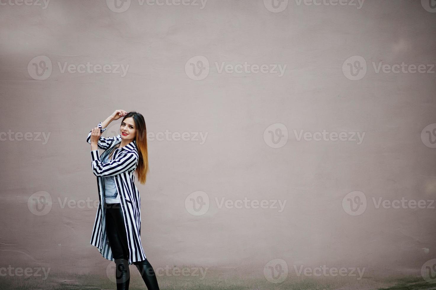 modischer frauenlook mit schwarz-weiß gestreifter anzugsjacke, lederhosen, die an der wand posieren. Konzept des Modemädchens. foto