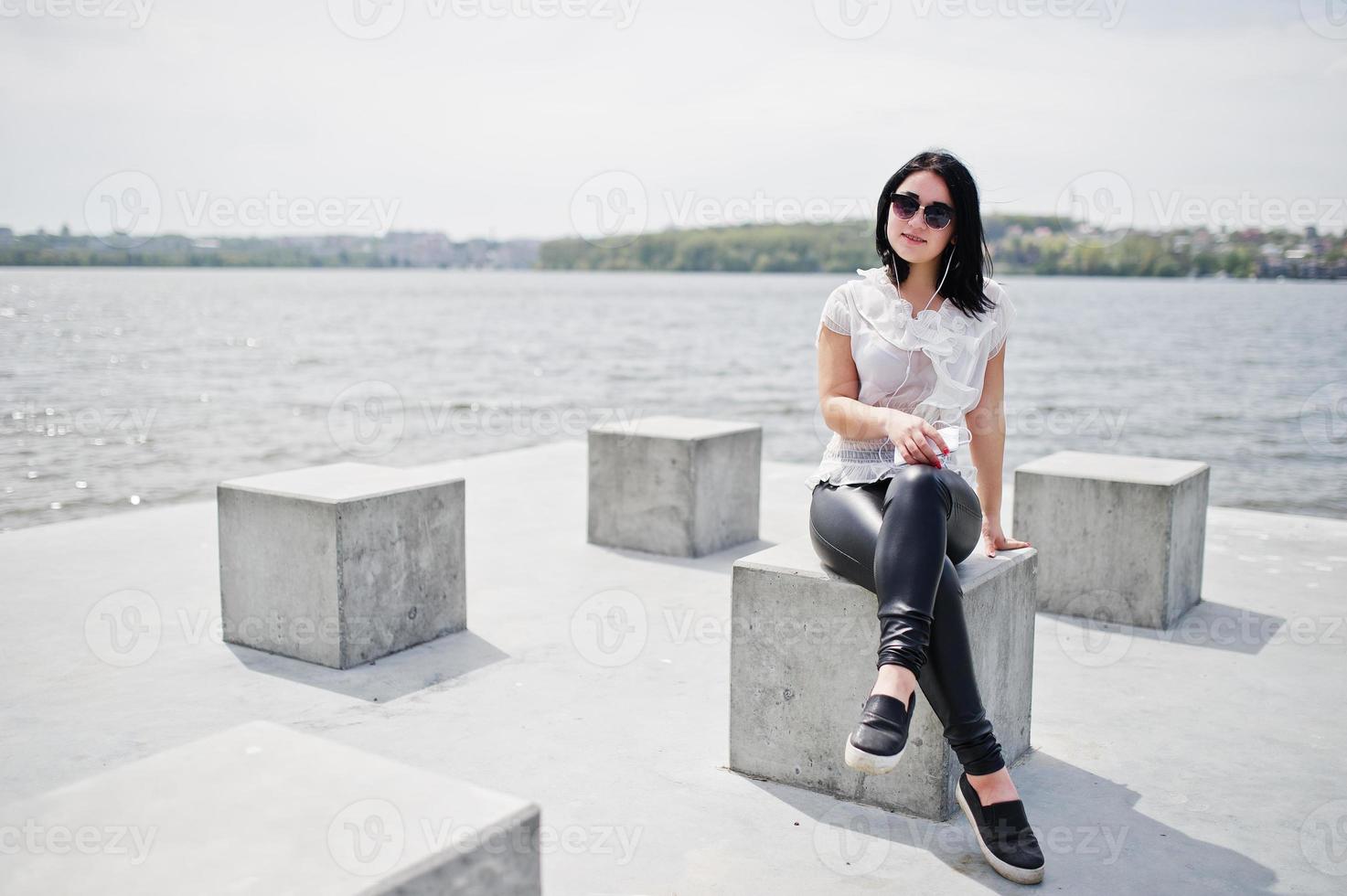 porträt eines brünetten mädchens, das musik über kopfhörer vom handy hört, lederhosen und weiße bluse für damen trägt, sonnenbrille, gegen steinwürfel am strand des sees. foto