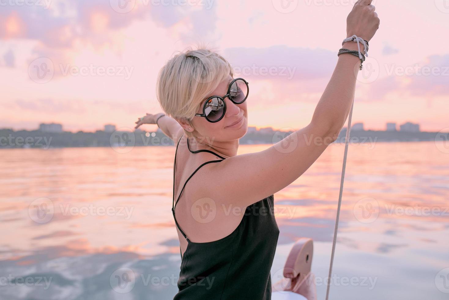 Schöne blonde glückliche Frau mit Sonnenbrille und langem schwarzem Kleid, die den Sonnenuntergang auf der Yacht im Meer genießt. reise-, abenteuer-, chill-, entspannungskonzept foto