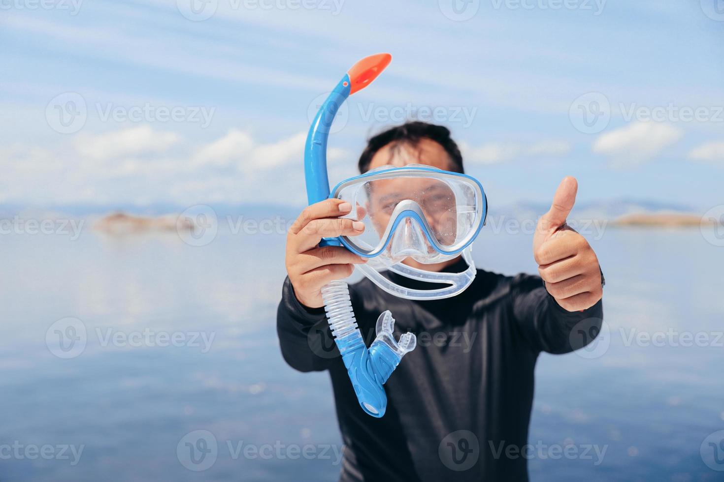 asiatischer mann im neoprenanzug, der schnorchelmaske hält und im sommerurlaub den daumen hochgibt foto