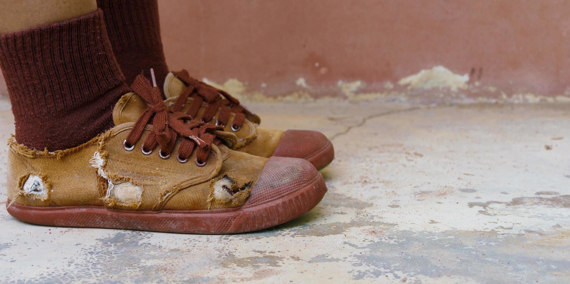 zerrissene Schülerschuhe, Armut ländlicher Schulkinder, die sich oft keine neuen Schuhe leisten können, Highschool-Schülerbeine mit zerrissenen Schuhen, schlechter Mangel an Unterrichtsmaterial, abgenutzte braune alte Turnschuhe foto