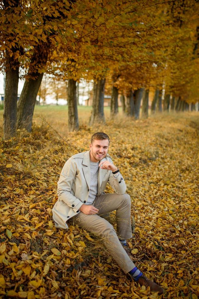 Porträt eines jungen Mannes in einem Mantel auf einem Hintergrund von Herbstbäumen. Ein Mann sitzt auf dem Boden. foto