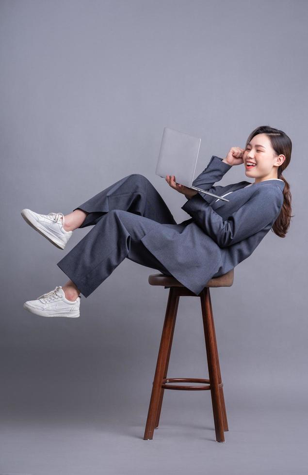 junge asiatische geschäftsfrau, die auf stuhl sitzt und laptop auf grauem hintergrund verwendet foto