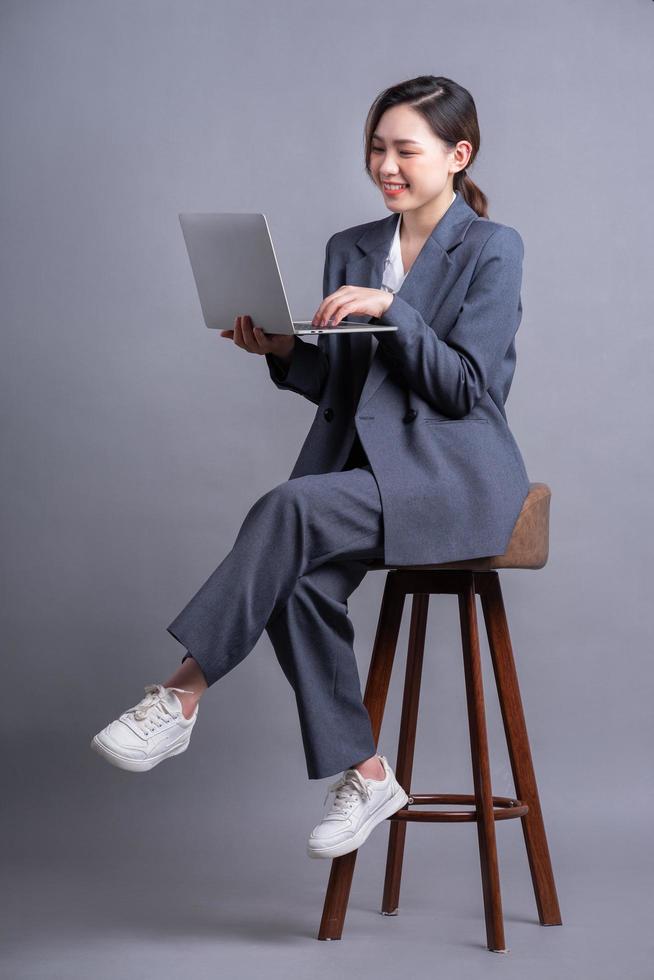 junge asiatische geschäftsfrau, die auf stuhl sitzt und laptop auf grauem hintergrund verwendet foto