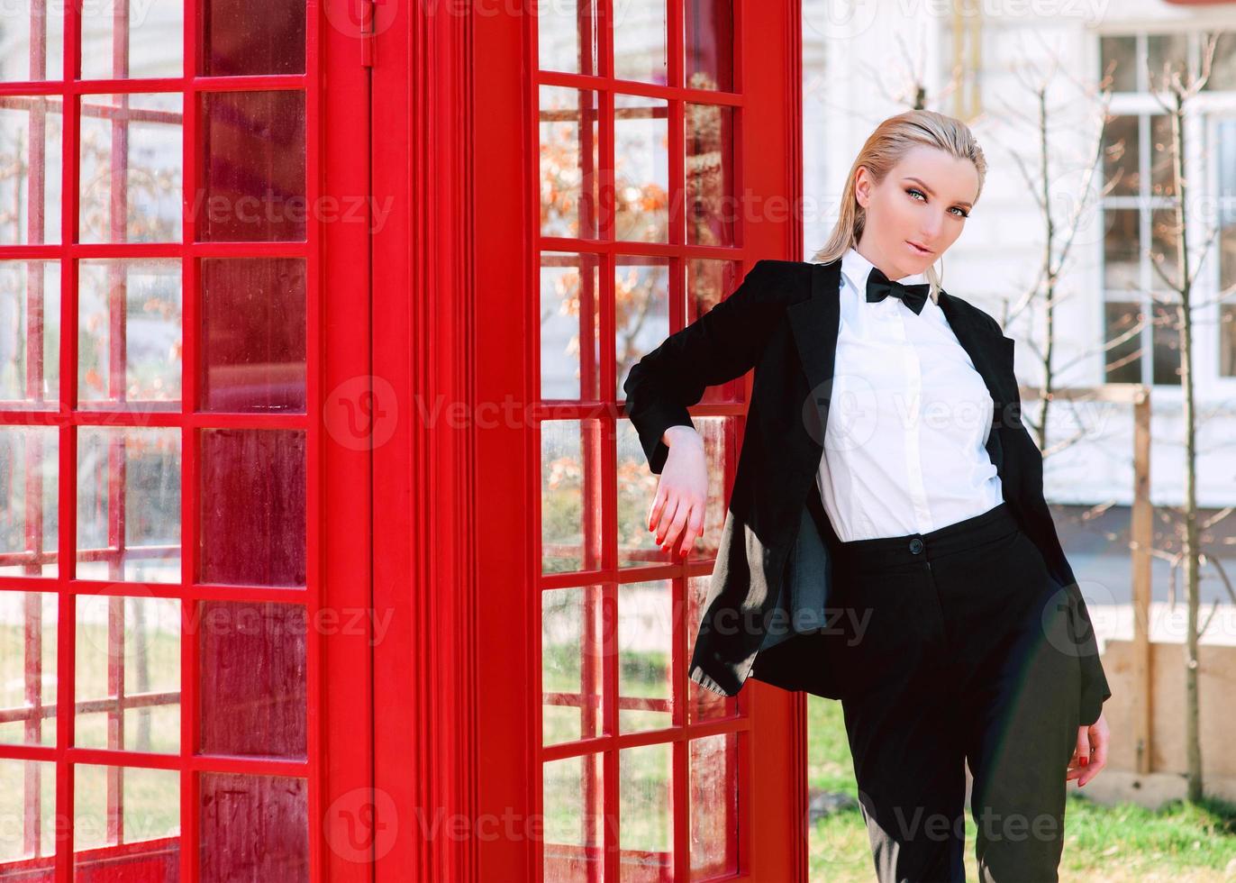 Porträt einer stilvollen, schönen blonden Frau im schwarzen Anzug eines Mannes in der Nähe einer roten Telefonzelle foto