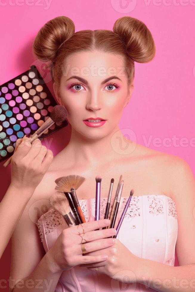 attraktive blonde Frau mit buntem Make-up mit Kosmetikpinseln und Schatten in ihr und den Händen des Make-up-Meisters foto