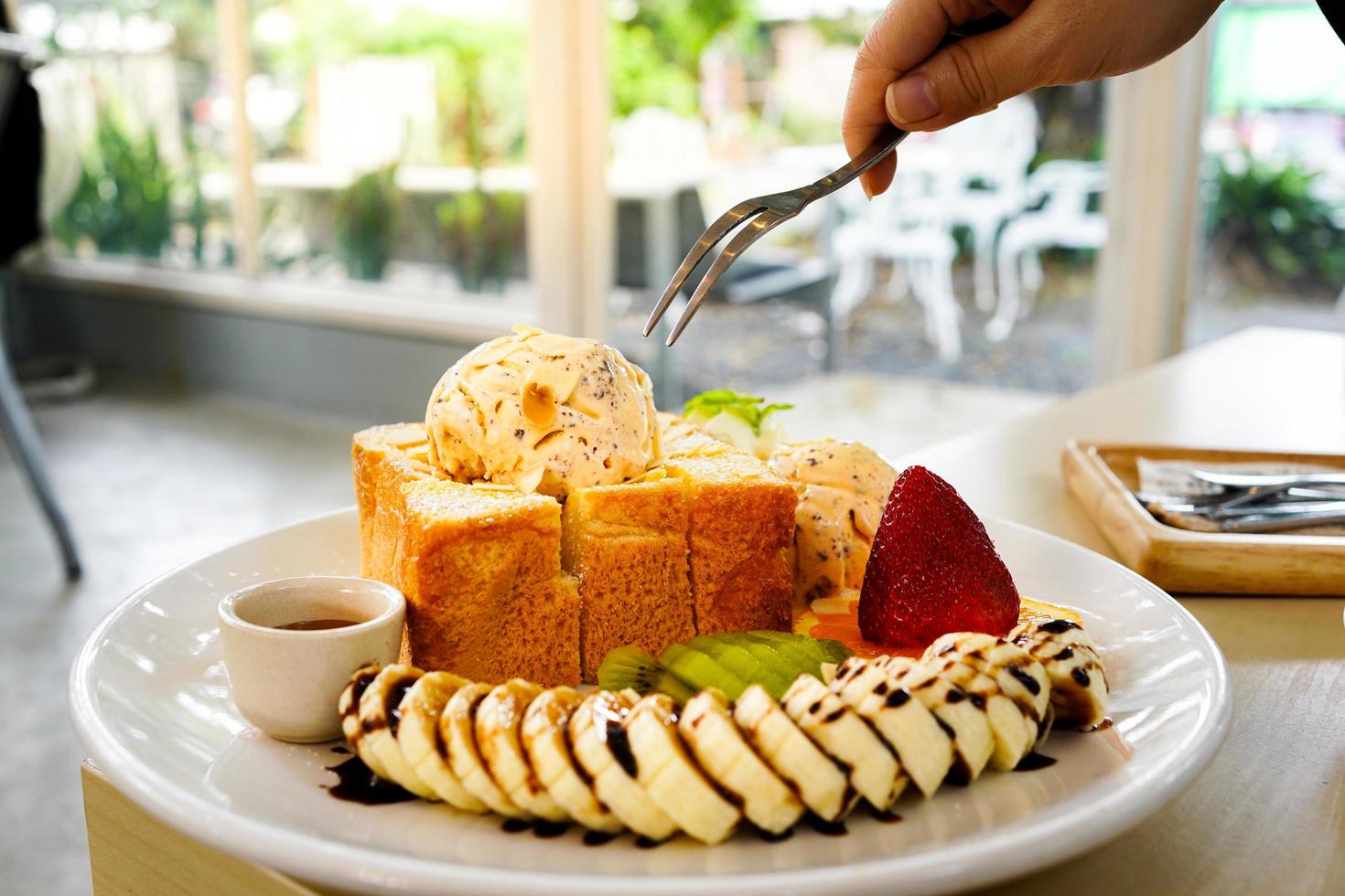 Selektiver Fokus, verwenden Sie eine Gabel, um Honigtoastbrot zu essen, das mit gemischten Früchten, Bananenscheiben, Eiscreme und Mandelscheiben und Honigsirup in einem weißen Teller serviert wird. foto