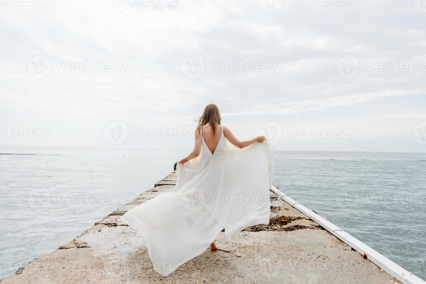 ein junges schönes mädchen in einem langen milchfarbenen kleid geht am strand und pier vor dem hintergrund des meeres entlang. foto