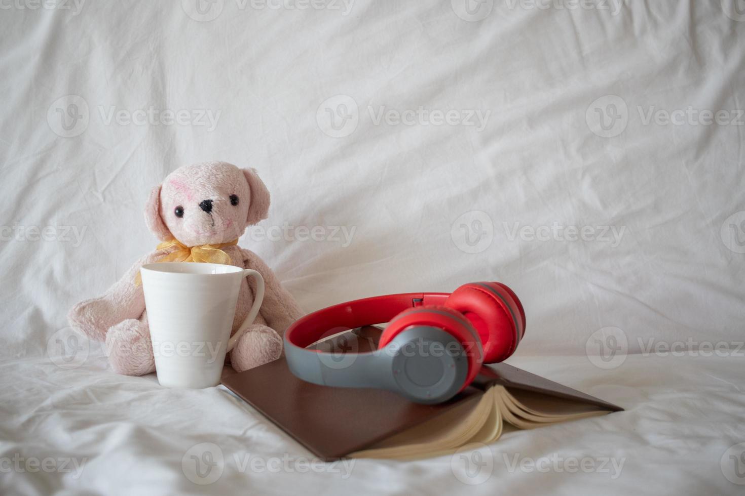 Ein roter Kopfhörer wird morgens auf ein Buch in der Nähe eines rosa Teddybären auf dem Bett gelegt, um sich bequem auszuruhen. ein kopfhörer und ein buch ruhen auf der weißen bettwäsche im schlafzimmer. foto