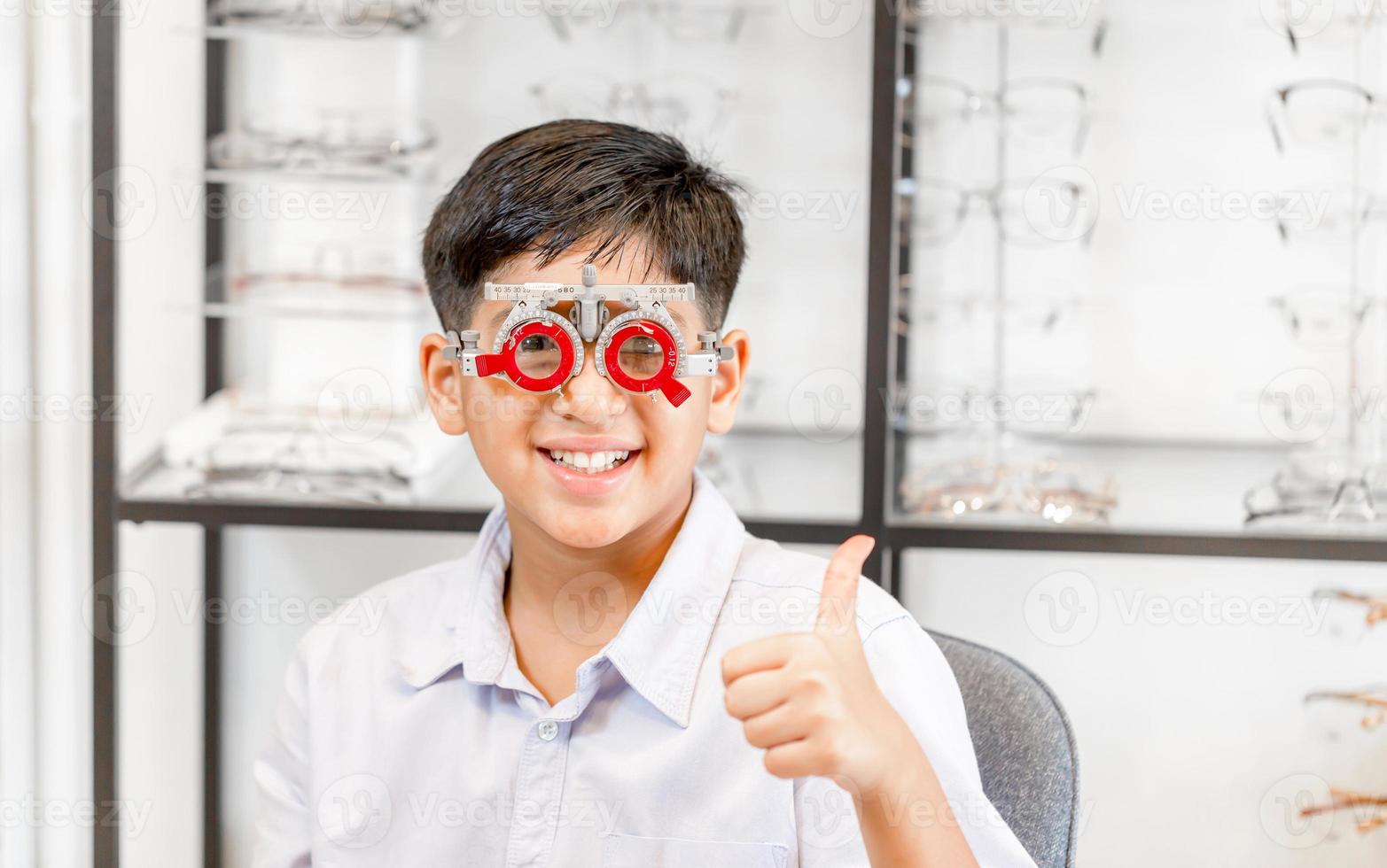 Lächelnder indisch-thailändischer Junge, der im Optikgeschäft eine Brille auswählt, Porträt eines Kindes, das im Optikgeschäft eine Augentestbrille trägt und Daumen nach oben zeigt foto
