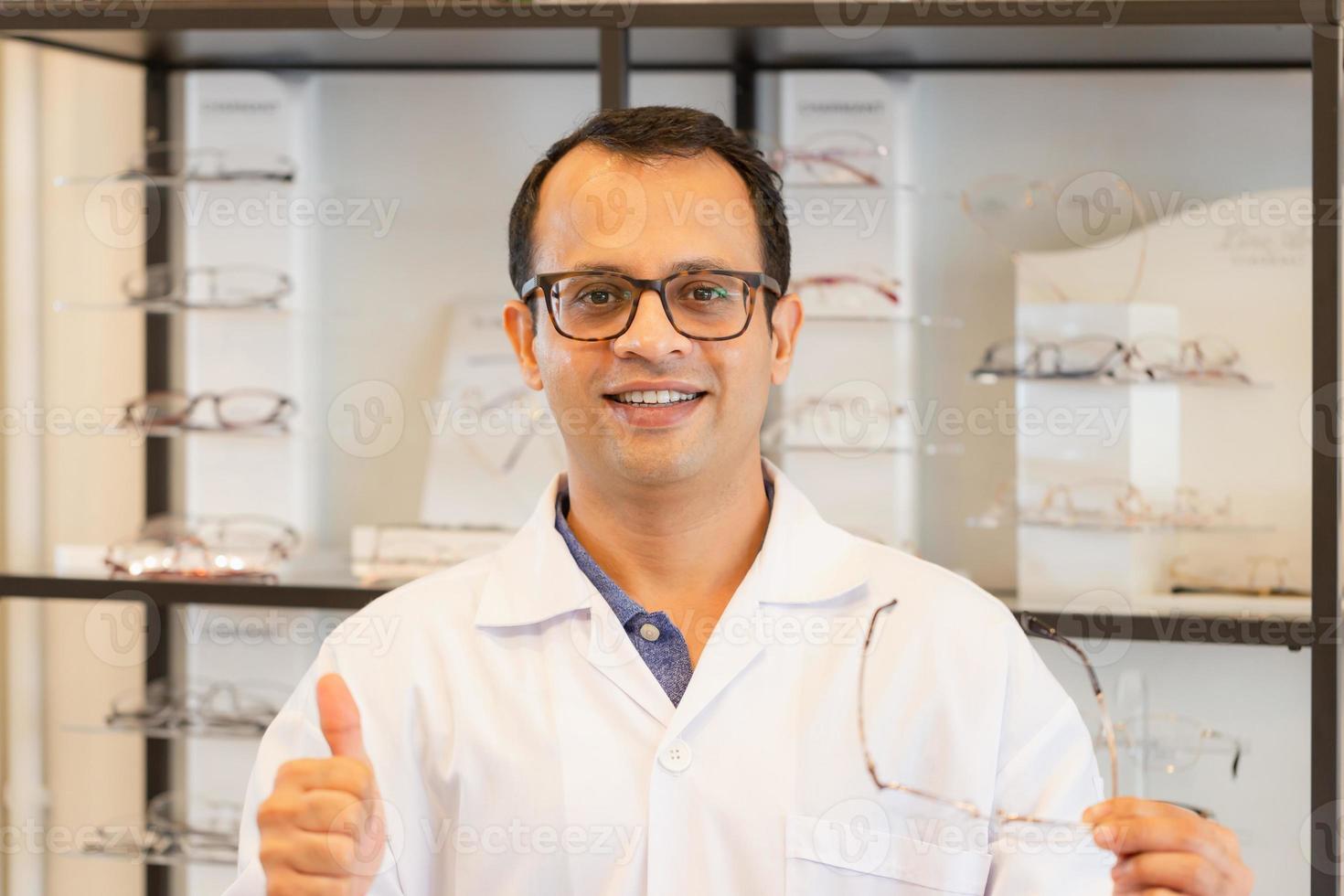 Fröhlicher Optometrist-Mann, der Brillen im Optikgeschäft hält, glücklicher indischer Mann, der Daumen nach oben im Optikgeschäft zeigt foto