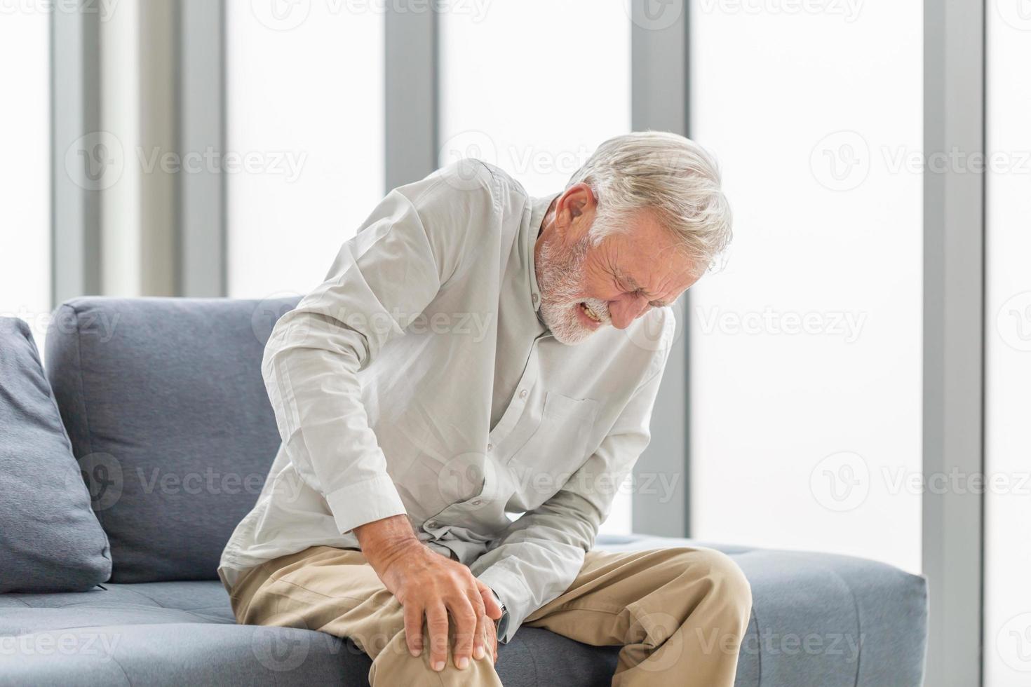 Alter Mann, der unter Knieschmerzen leidet, sitzt auf dem Sofa im Wohnzimmer, älterer Mann, der unter Knieschmerzen leidet, während er auf dem Sofa sitzt foto