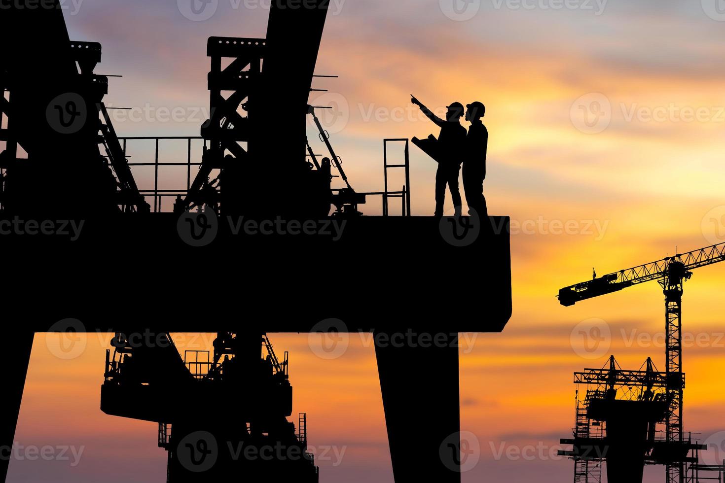 Silhouette von Ingenieuren und Arbeitern, die das Projekt auf Baustellenhintergrund der schweren Infrastruktur, Baustelle bei Sonnenuntergang in der Abendzeit überprüfen. foto