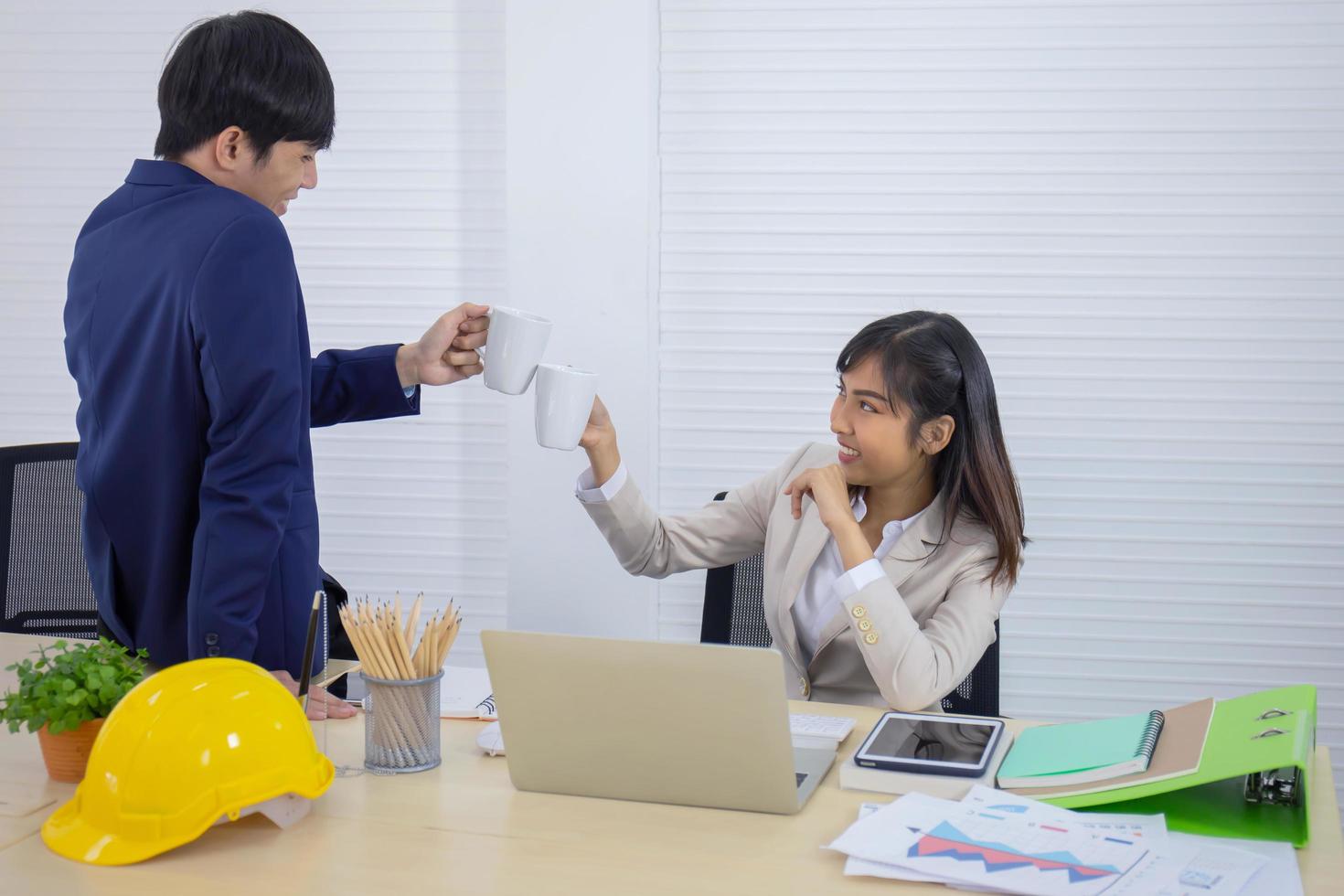 eine junge, professionelle junge asiatische geschäftsfrau sitzt an ihrem schreibtisch und stößt mit einem männlichen büroangestellten, der zusammenarbeitet, eine tasse kaffee an. foto