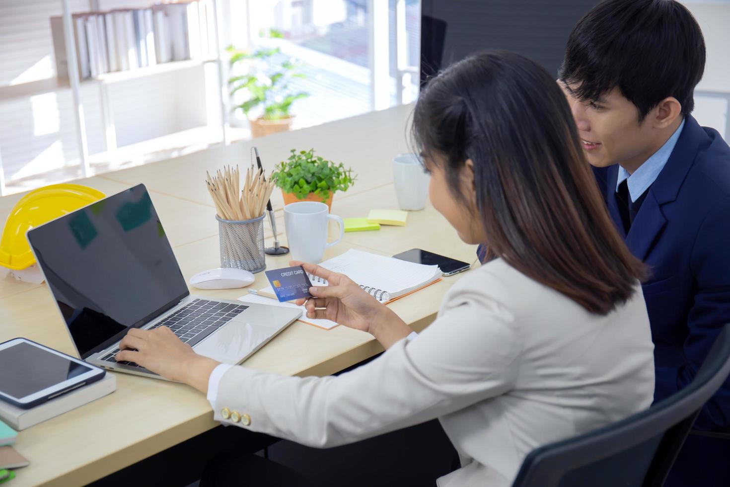 eine junge, professionelle asiatische geschäftsfrau, die mit einem laptop und einer kreditkarte online bezahlt, während sie an ihrem schreibtisch sitzt, mit einem männlichen freund neben ihm. foto