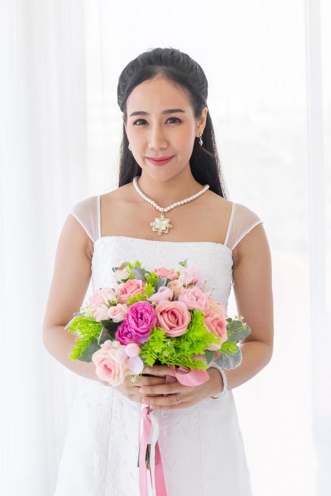 eine asiatische braut in einem weißen hochzeitskleid steht strahlend lächelnd in einer hand, die einen schönen blumenstrauß hält. foto