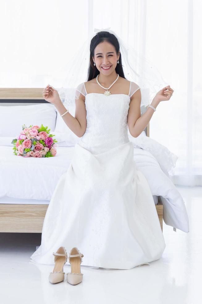 eine asiatische braut in einem weißen hochzeitskleid saß auf dem bett und lächelte strahlend, mit einem blumenstrauß neben ihr. foto