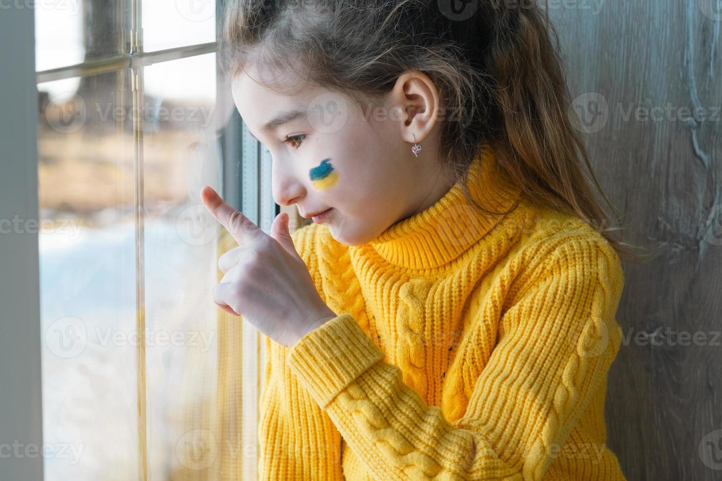 ein trauriges kind schaut auf das fenster mit der flagge der ukraine auf die wange gemalt, sorgen und angst. humanitäre Hilfe für Kinder, Weltfrieden, Sicherheit. foto