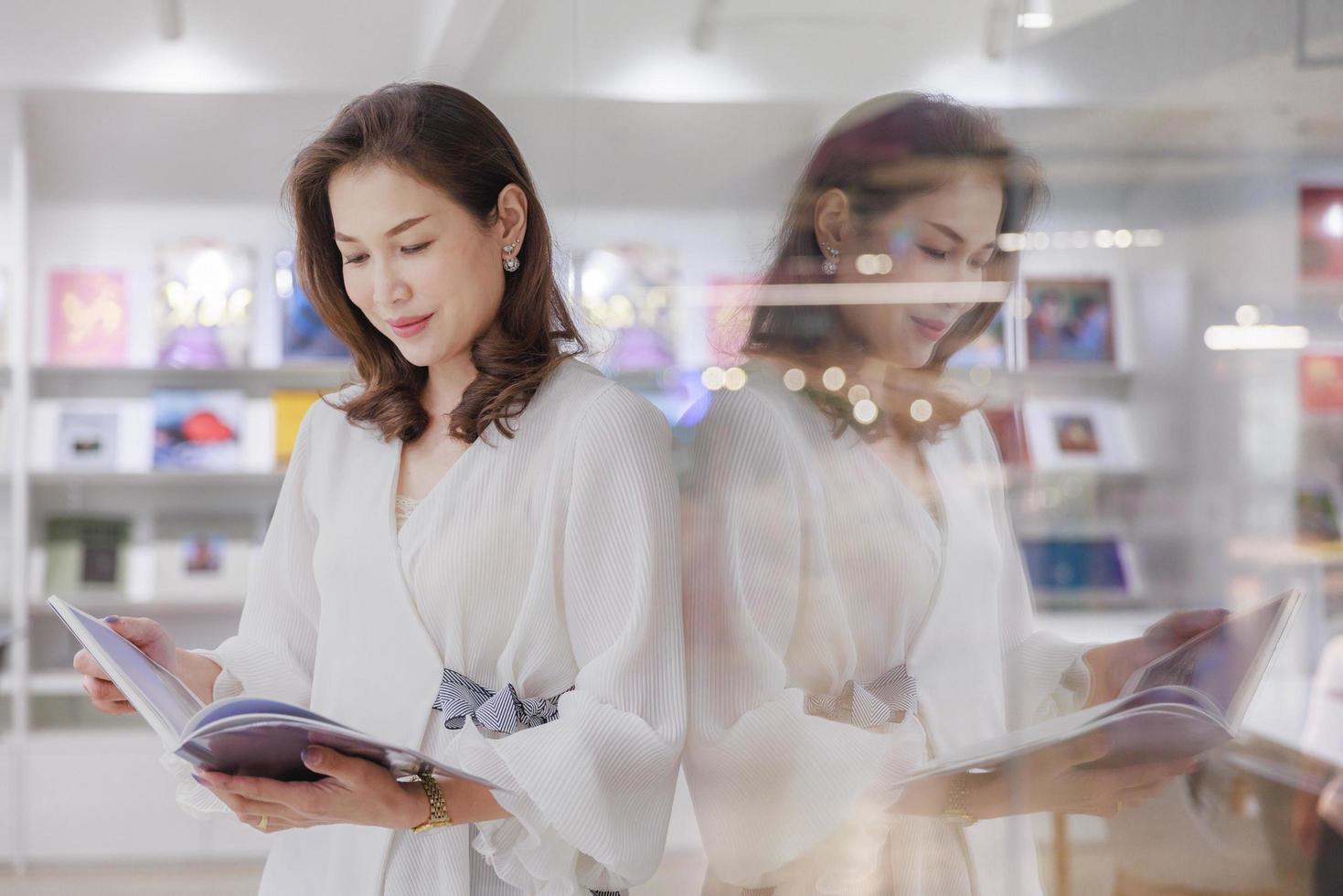 Porträt einer hübschen, fröhlichen Frau, die in der Bibliothek steht und ein Buch liest und sich glücklich an die Glaswand lehnt, sich konzentriert und mit Reflexion im klaren Spiegel konzentriert foto