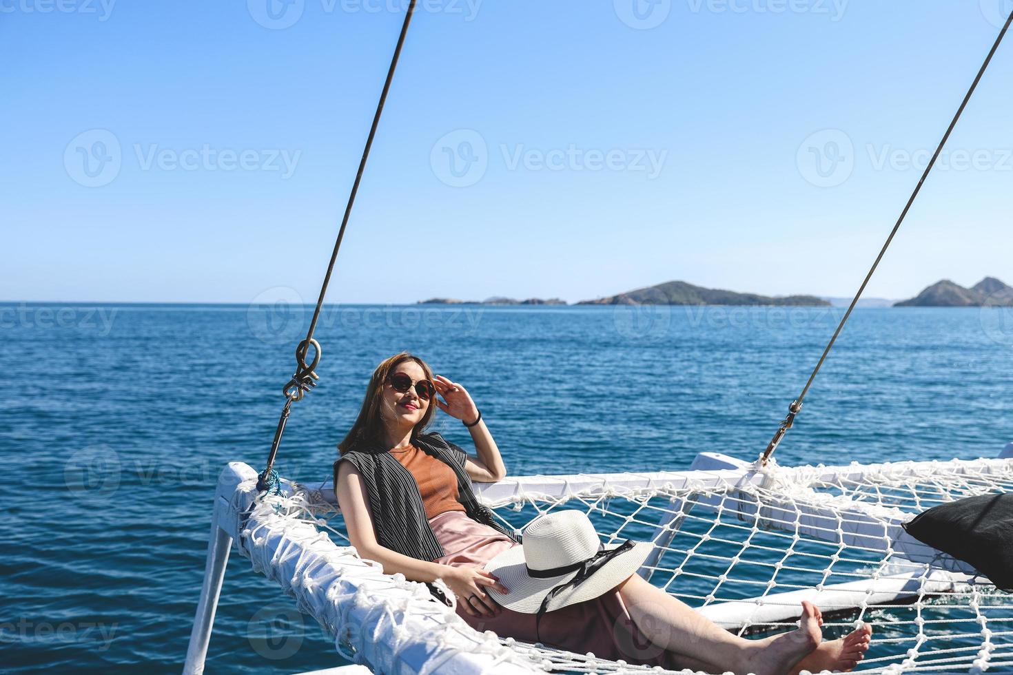 Lächelnde asiatische Frau mit Sonnenbrille, die sich auf dem Katamaran-Netz entspannt und die Sommerferien genießt foto