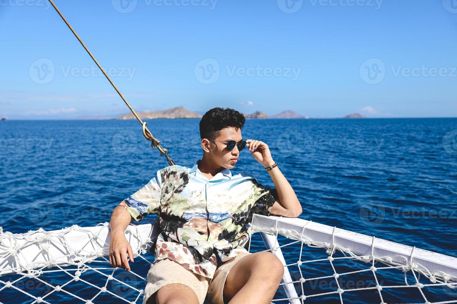 asiatischer mann mit sonnenbrille, der auf einem katamarannetz sitzt und sich entspannt, während er die seelandschaft genießt foto