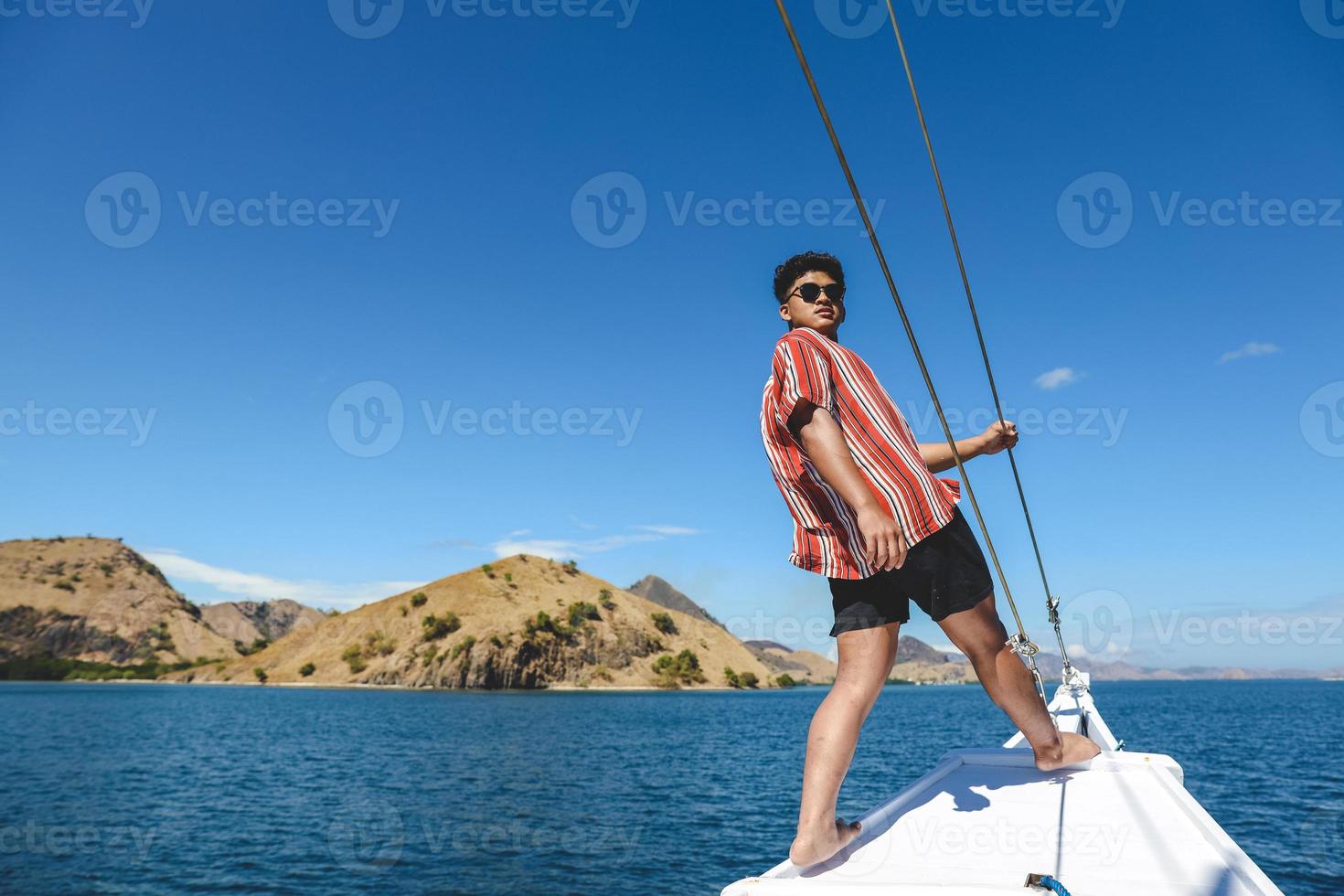 asiatischer mann mit sonnenbrille, der am rand des bootes mit seelandschaft und hügeln bei labuan bajo posiert foto