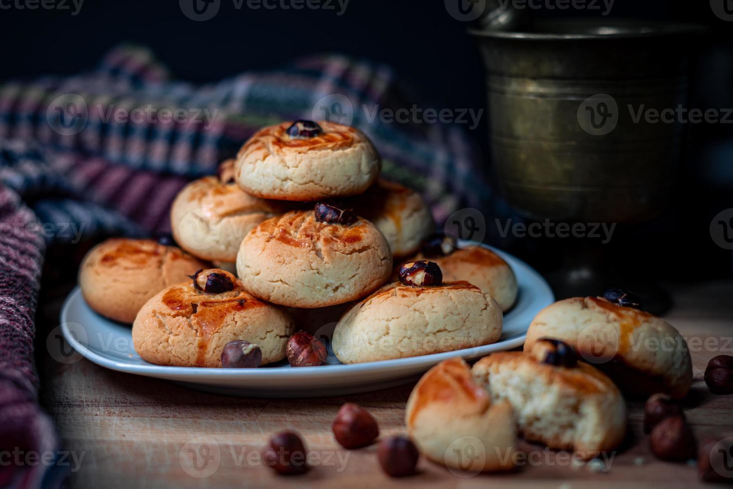 Knusprige Kekse, die in einem Teller auf einem Holzbrett mit Haselnusskernstößel und Tischdecke gestapelt sind foto