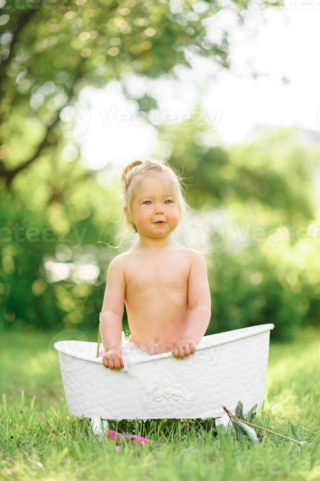 glückliches Kleinkind nimmt ein Milchbad mit Blütenblättern. kleines Mädchen in einem Milchbad auf grünem Hintergrund. Blumensträuße aus rosa Pfingstrosen. baby baden. Hygiene und Pflege für Kleinkinder. foto