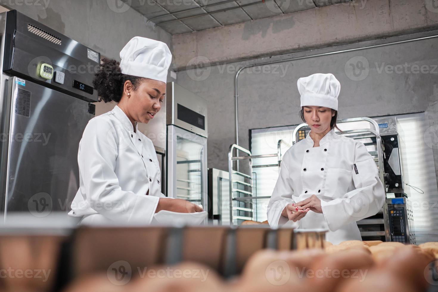 Zwei professionelle Köchinnen in weißen Kochuniformen und Schürzen kneten Teig und Eier, bereiten Brot, Kekse und frische Backwaren zu und backen in einer Edelstahlküche eines Restaurants im Ofen. foto