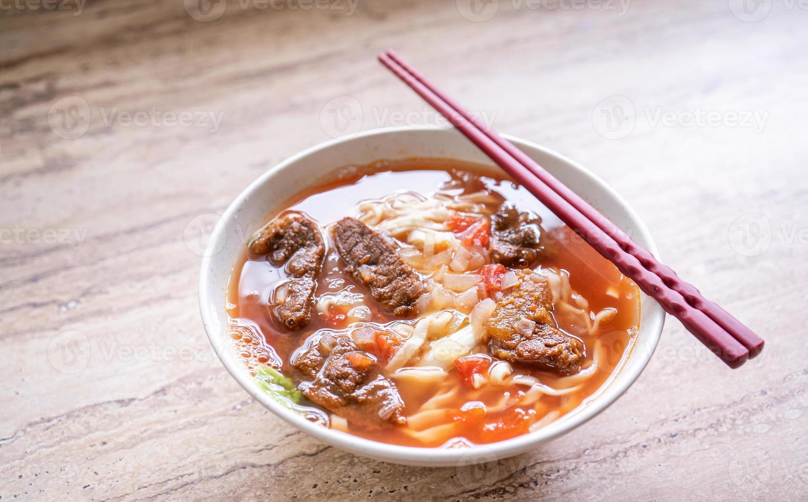 Rindfleisch-Nudel-Ramen-Mahlzeit mit Tomatensauce-Brühe in Schüssel auf hellem Holztisch, berühmtes Essen im chinesischen Stil in Taiwan, Nahaufnahme, Draufsicht, Kopierraum foto