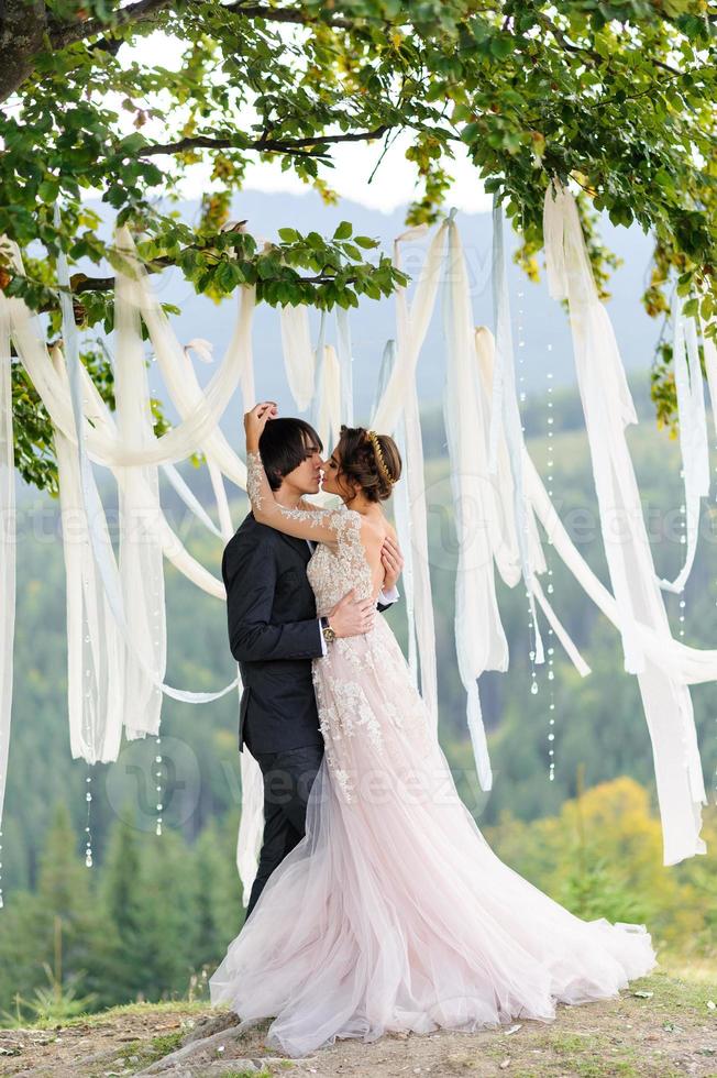 Braut und Bräutigam umarmen sich unter einer alten Eiche. Hochzeitsfotoshooting in den Bergen. daneben ist Dekor für die Zeremonie vorbereitet. foto