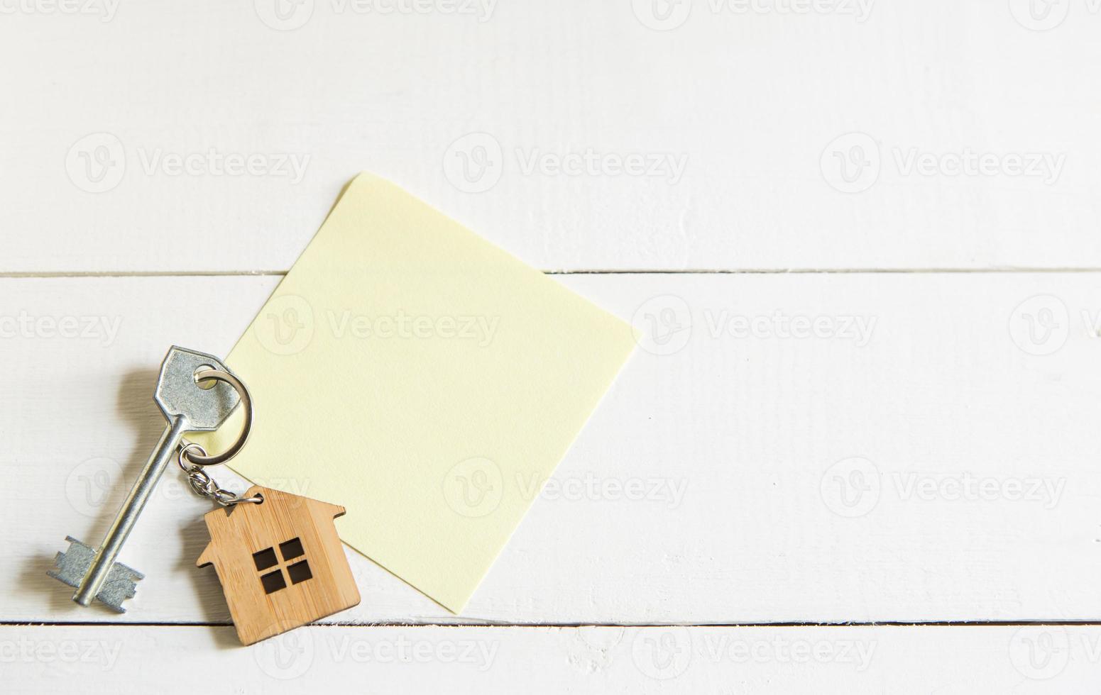 Schlüsselanhänger in Form eines Holzhauses mit Schlüssel auf weißem Hintergrund mit einem quadratischen Blatt für Notizen. Bau, Design, Projekt, Umzug in ein neues Haus, Hypothek, Miete und Kauf von Immobilien. Platz kopieren foto