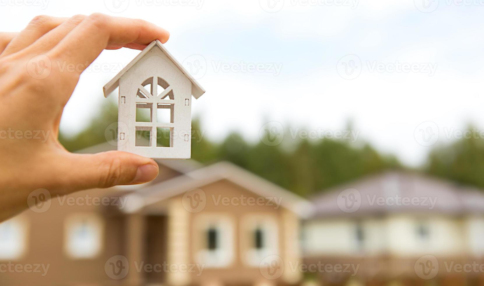 Modell eines weißen Holzhauses in der Hand vor dem Hintergrund der Hütten im Dorf. Traum vom Eigenheim, Bauen, Gestalten, Umzug in ein neues Haus, Hypothek, Miete und Kauf von Immobilien. Platz kopieren foto