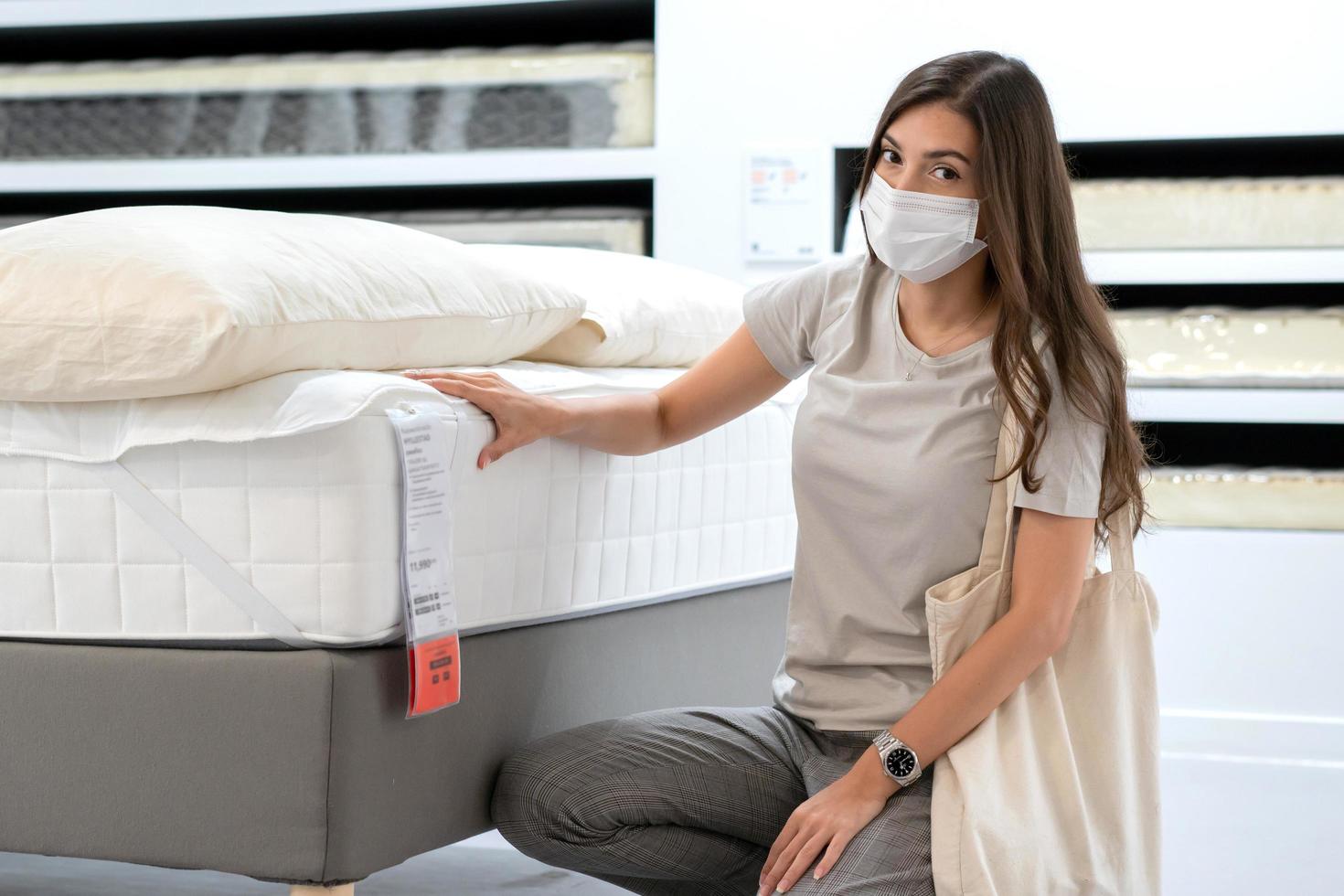 Frau mit Schutzmaske, die eine Bettmatratze für den Umzug auswählt oder ein neues Zimmer dekoriert. neues normales einkaufen während der coronavirus-pandemie. foto