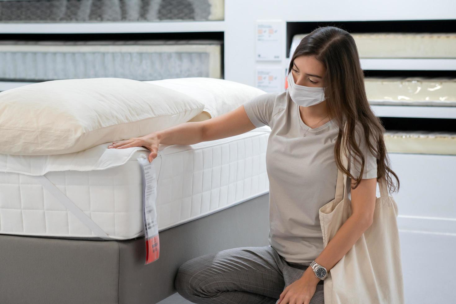 Frau mit Schutzmaske, die eine Bettmatratze für den Umzug auswählt oder ein neues Zimmer dekoriert. neues normales einkaufen während der coronavirus-pandemie. foto